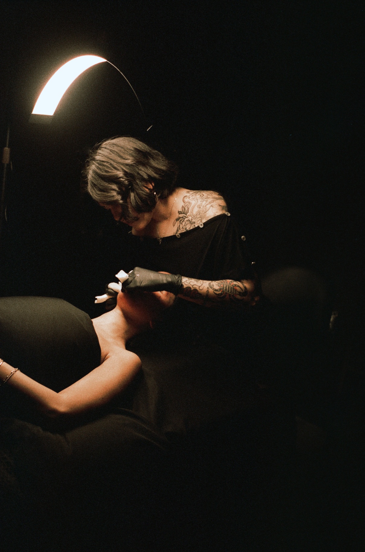Tattooed esthetician performing precise detail work under an arched task lamp